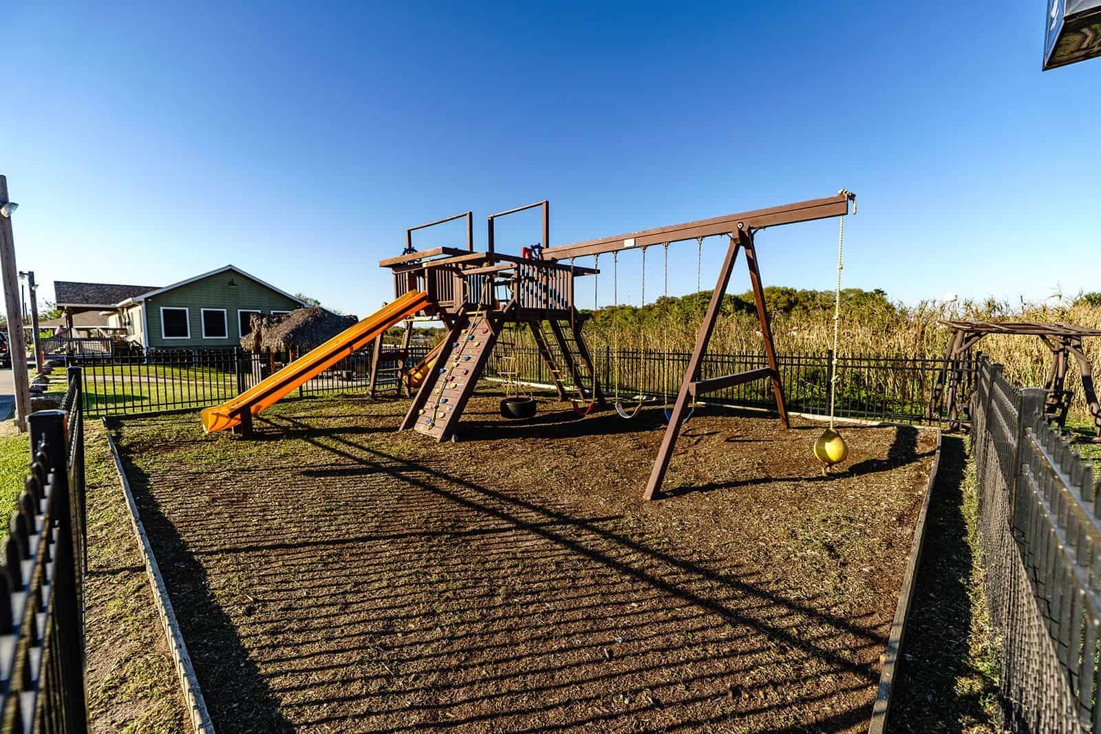 Fenced playground with wooden play structure, orange slide, swings, and climbing wall
